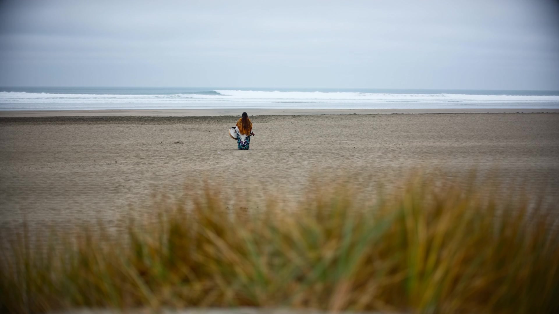 Amikaeyla on a wide Pacific beach with a frame drum, dune grass in the foreground, ocean waves in the distance