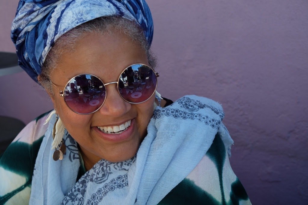 Amikaeyla wearing a blue tie-dyed turban and round purple sunglasses, smiling against a lavender wall