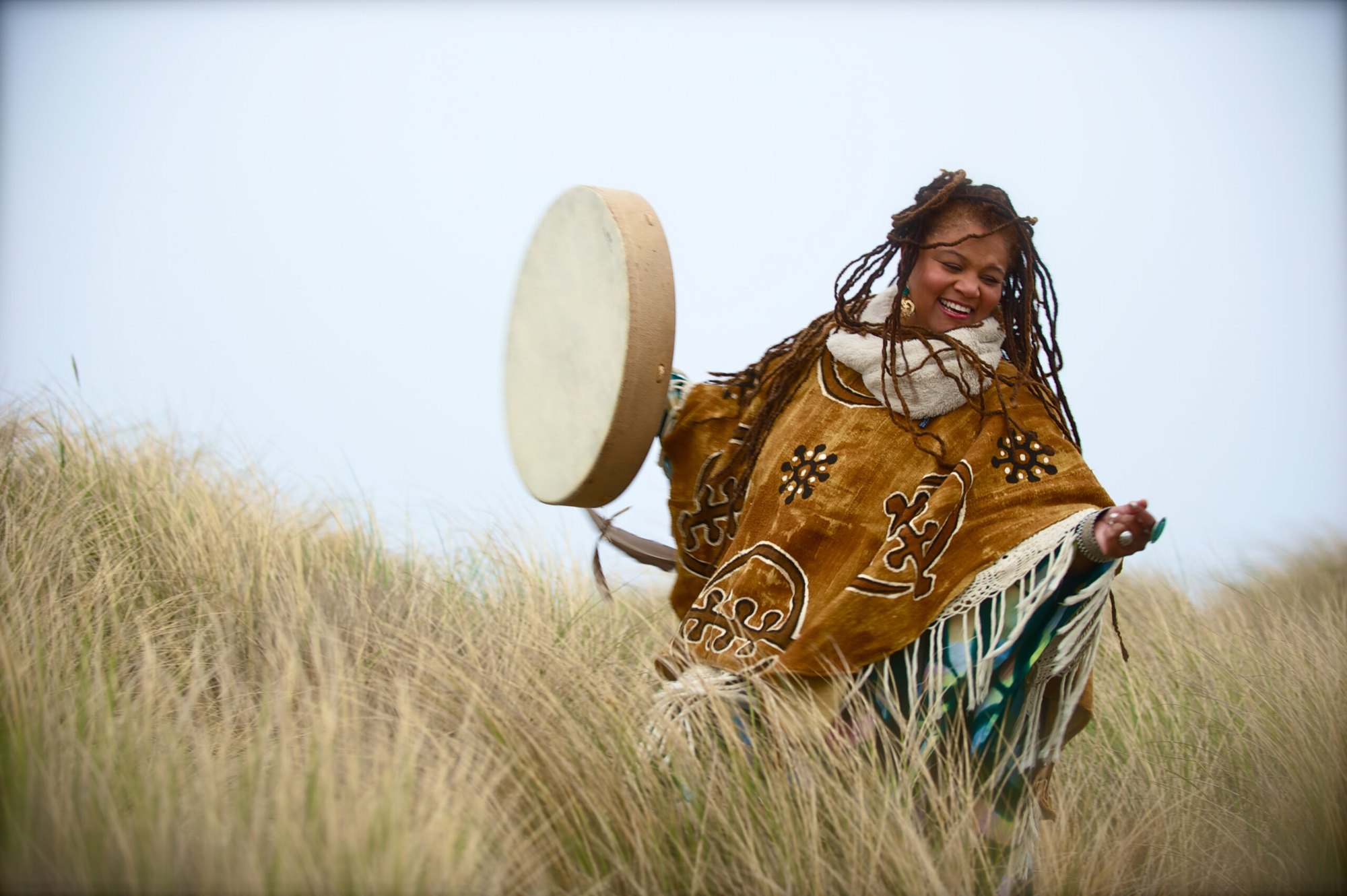 Amikaeyla in a gold mudcloth poncho, frame drum mid-toss, laughing in dune grass on a Pacific beach