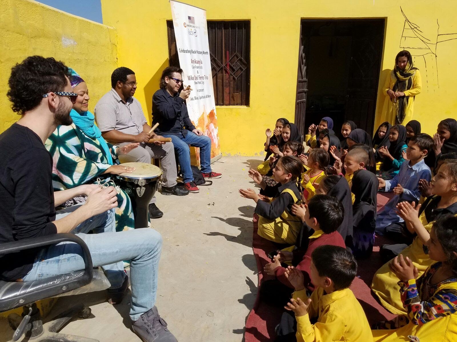 Amikaeyla and her band performing for schoolchildren in Pakistan, seated against a yellow wall