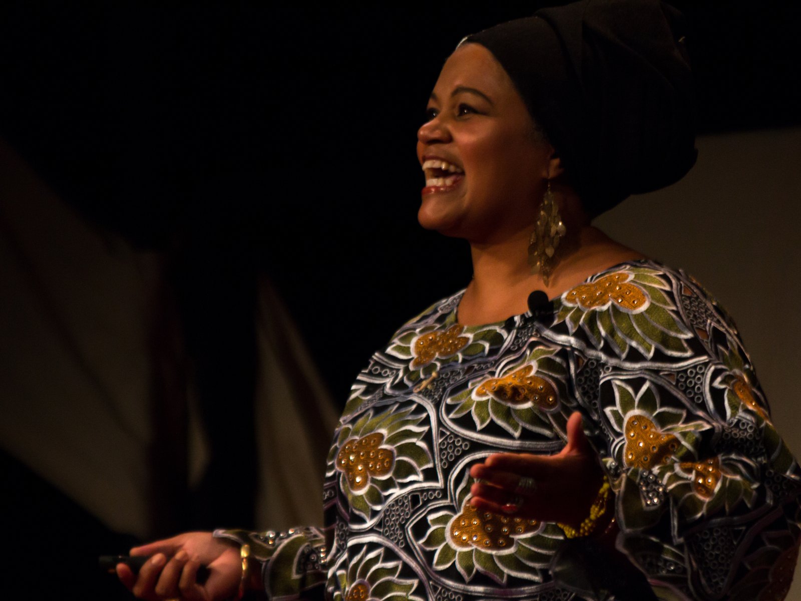 Amikaeyla mid-laugh on stage in a green-and-gold patterned gown and black headwrap against a dark backdrop