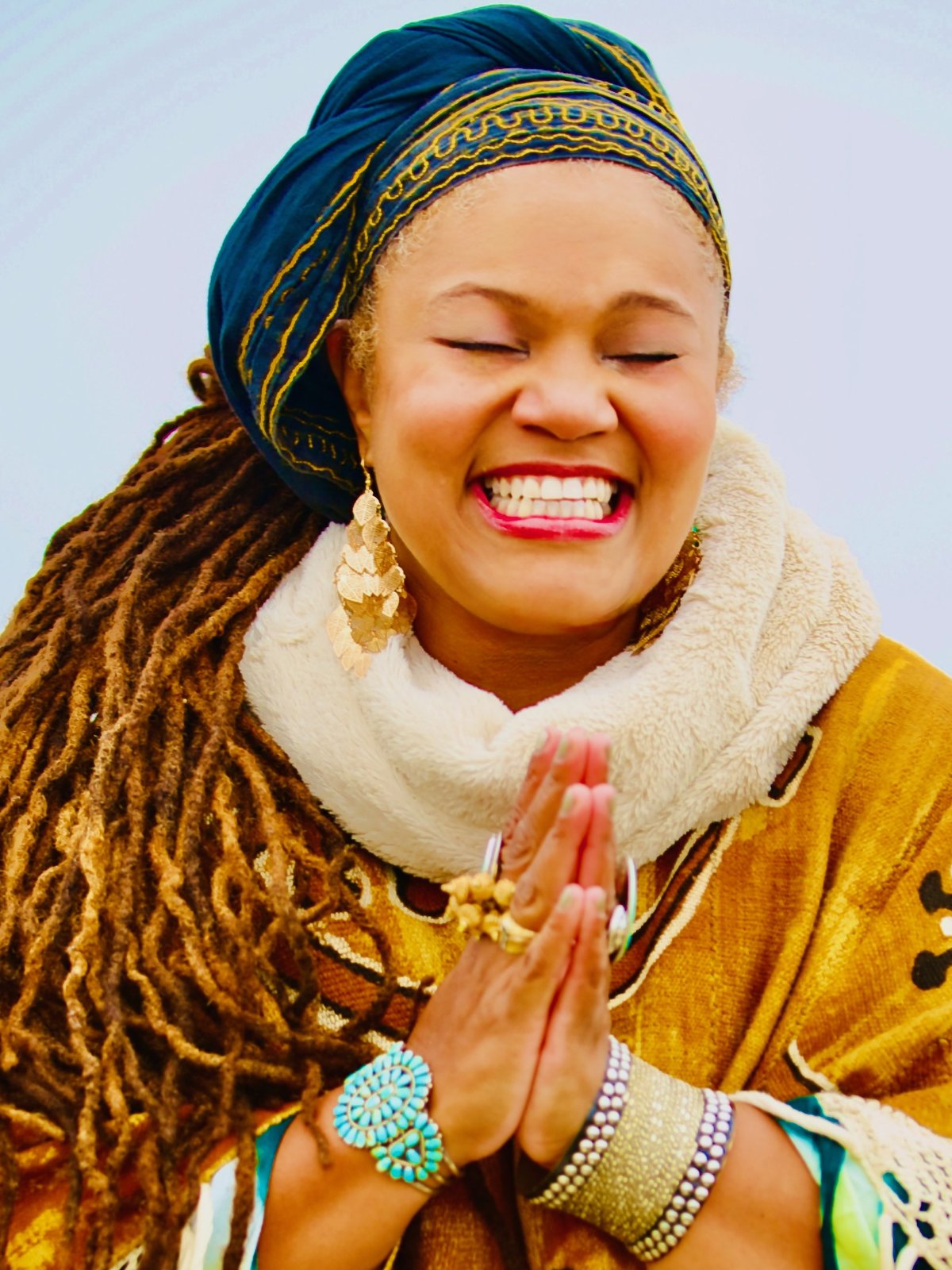 Amikaeyla Gaston smiling with eyes closed, hands pressed together in gratitude, in an indigo-and-gold headwrap and honey robe against a soft dawn-blue sky